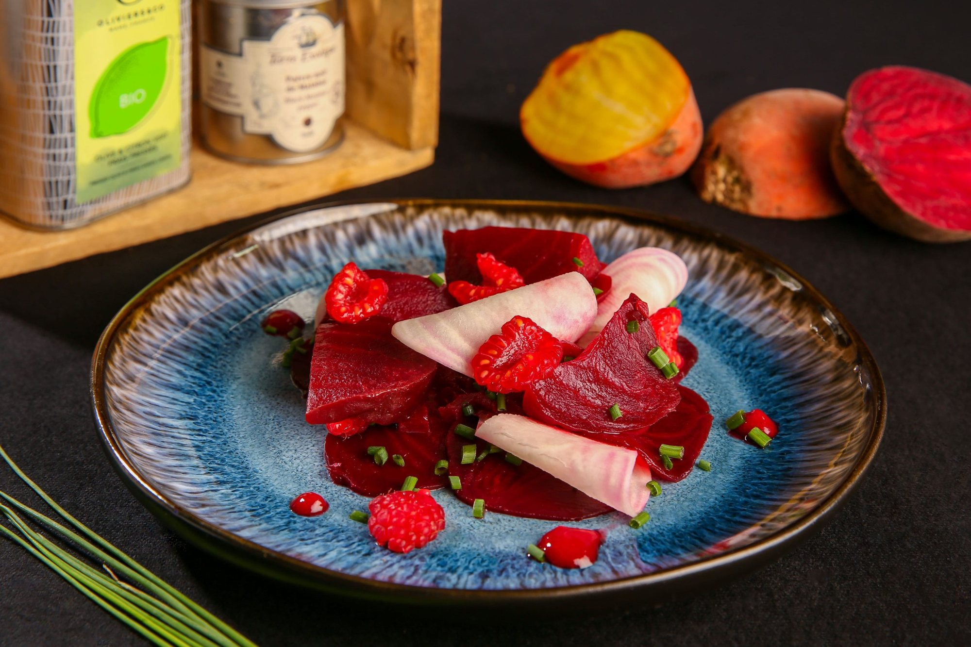 Une assiette en céramique bleue garnie de tranches de betterave, de framboises et de ciboulette, posée devant des betteraves entières et des bidons d'huile sur un support en bois.