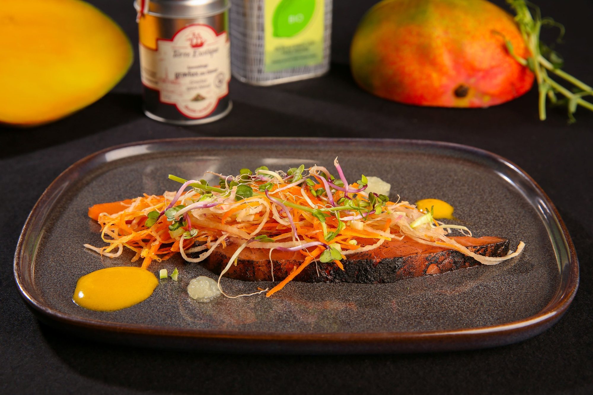 Une assiette grise carrée garnie d'une tranche de saumon mariné, de julienne de légumes et de pousses, entourée de mangues et de pots d'épices sur une nappe noire.