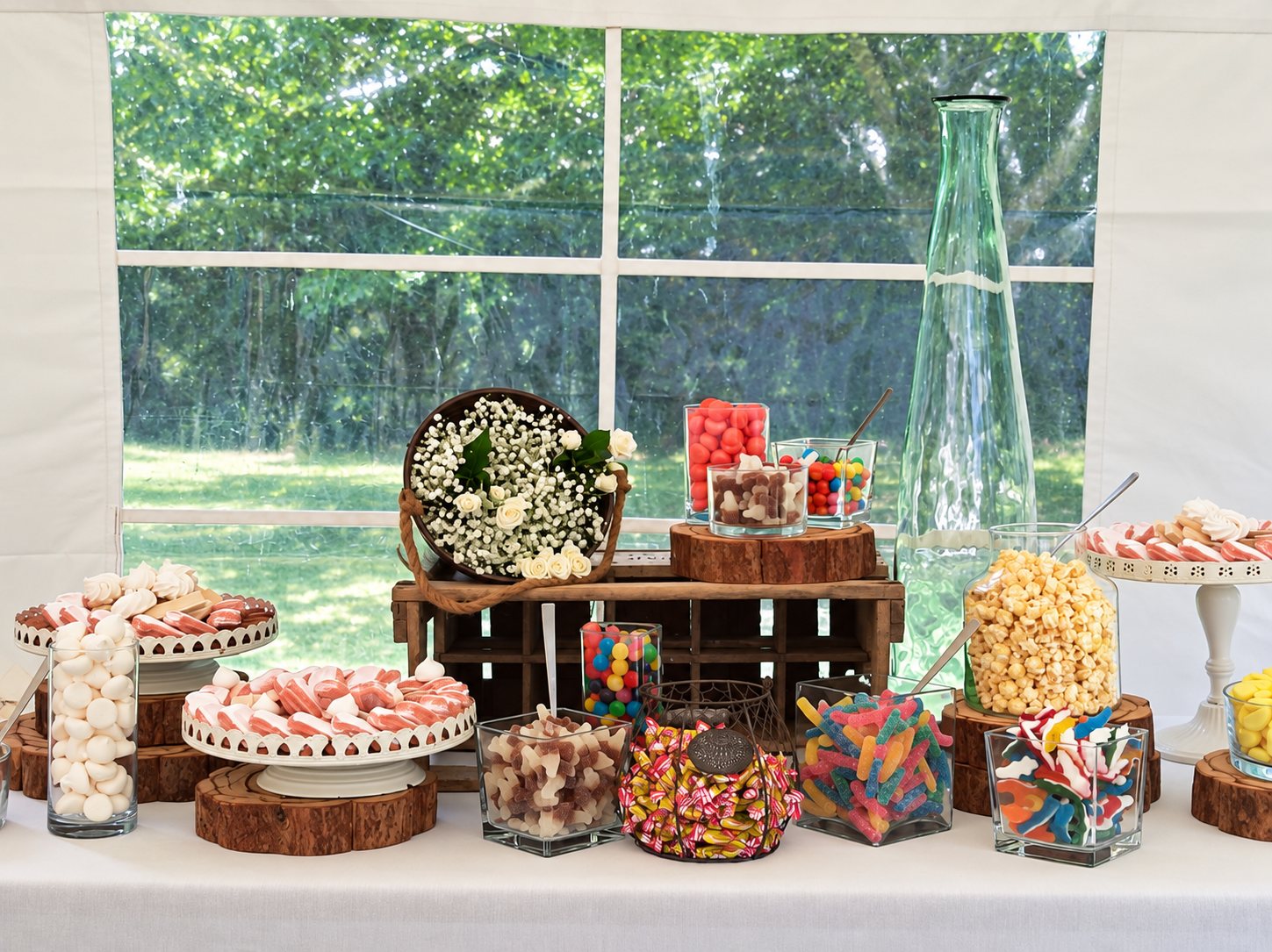 Table de bonbons décorée de gypsophile, de rondins de bois et de bocaux en verre remplis de confiseries, de pop-corn et de meringues, installée sous une tente blanche.
