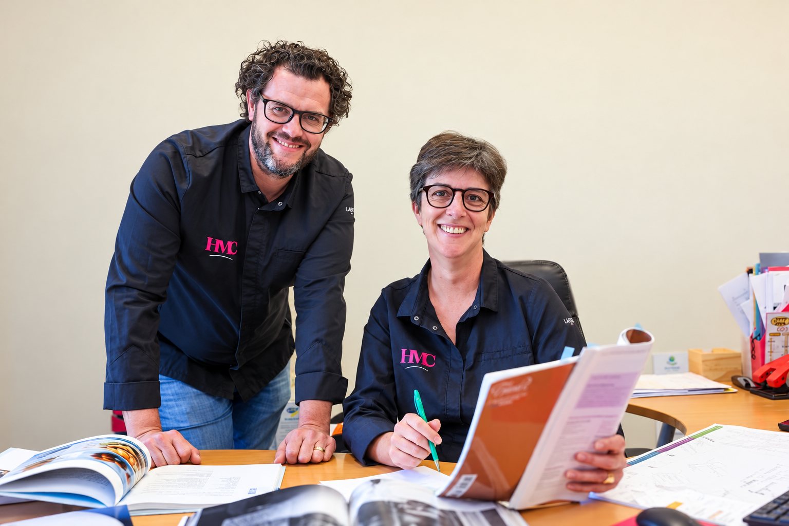 Un homme et une femme portant des vestes noires siglées HMC sourient à la caméra à un bureau couvert de catalogues et de documents.