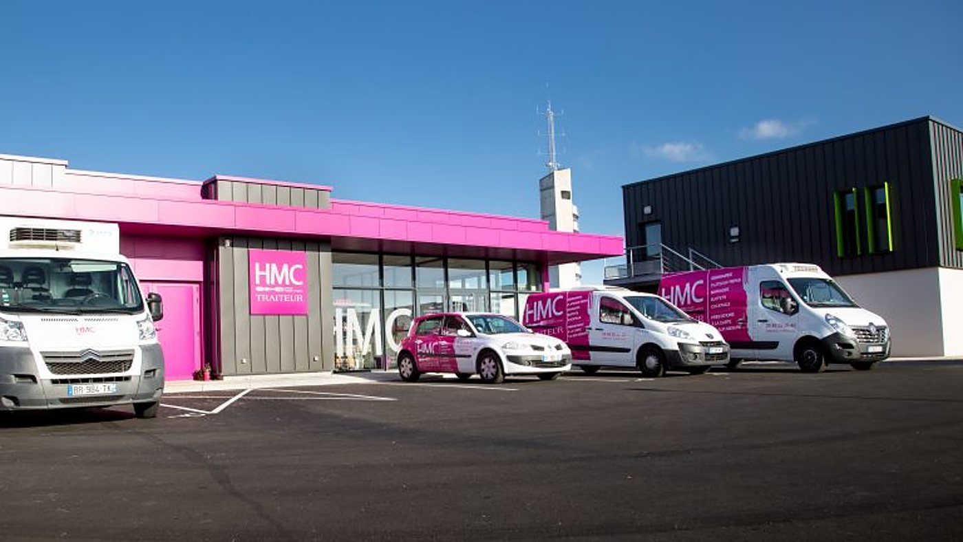 Devant le bâtiment rose du traiteur HMC, deux camionnettes blanches et roses, une voiture assortie et un autre utilitaire sont garés sur le parking.