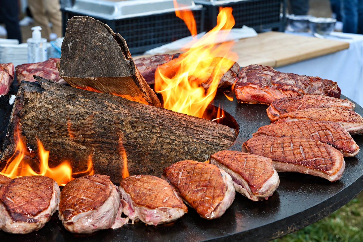 Des magrets de canard quadrillés et des pièces de viande cuisent sur une grande plancha ronde en métal autour de bûches en flammes, en plein air.