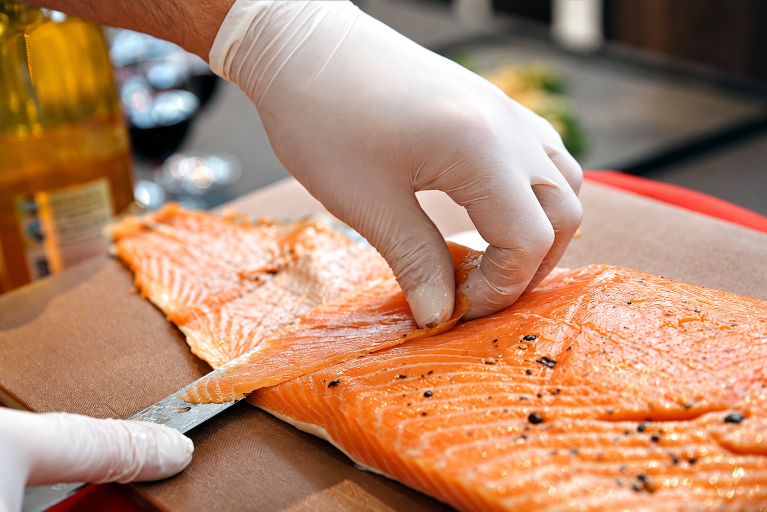 Des mains portant des gants blancs retirent la peau d'un filet de saumon poivré posé sur une planche à découper, un couteau à côté.