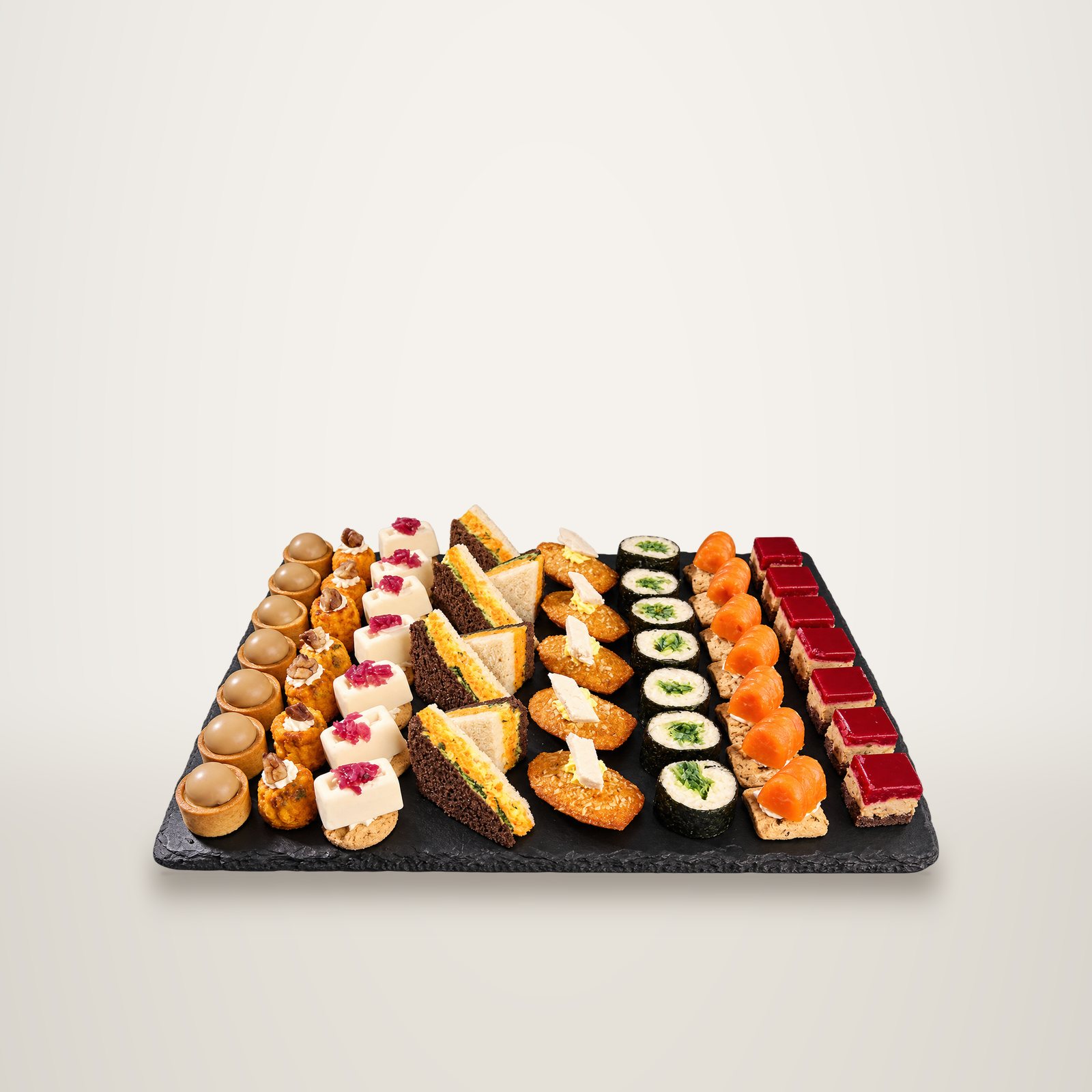 Une ardoise noire présente des rangées de petits fours salés et sucrés : tartelettes, sandwichs, madeleines, makis, saumon et bouchées rouges, sur fond noir.