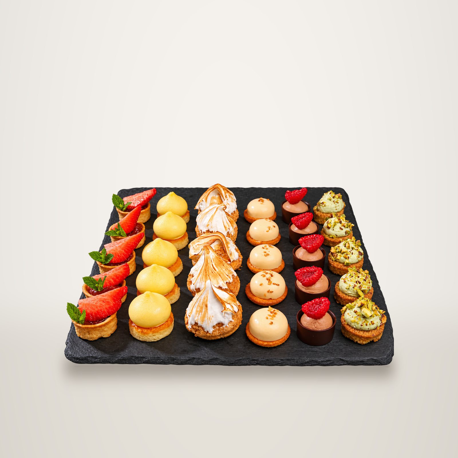 Un plateau d'ardoise présente des petites pâtisseries alignées en colonnes : tartelettes aux fraises, choux meringués, mousses à la framboise et tartelettes à la pistache.
