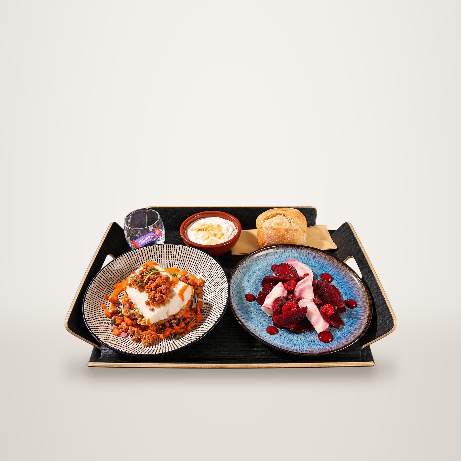 Plateau noir présentant une assiette de poisson blanc au chorizo, une assiette de betteraves et framboises, un petit pain, une crème aux amandes et un verre avec un chocolat Milka.
