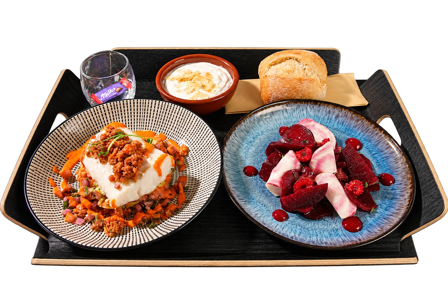 Plateau noir présentant une assiette de poisson blanc au chorizo, une assiette de betteraves et framboises, un petit pain, une crème aux amandes et un verre avec un chocolat Milka.