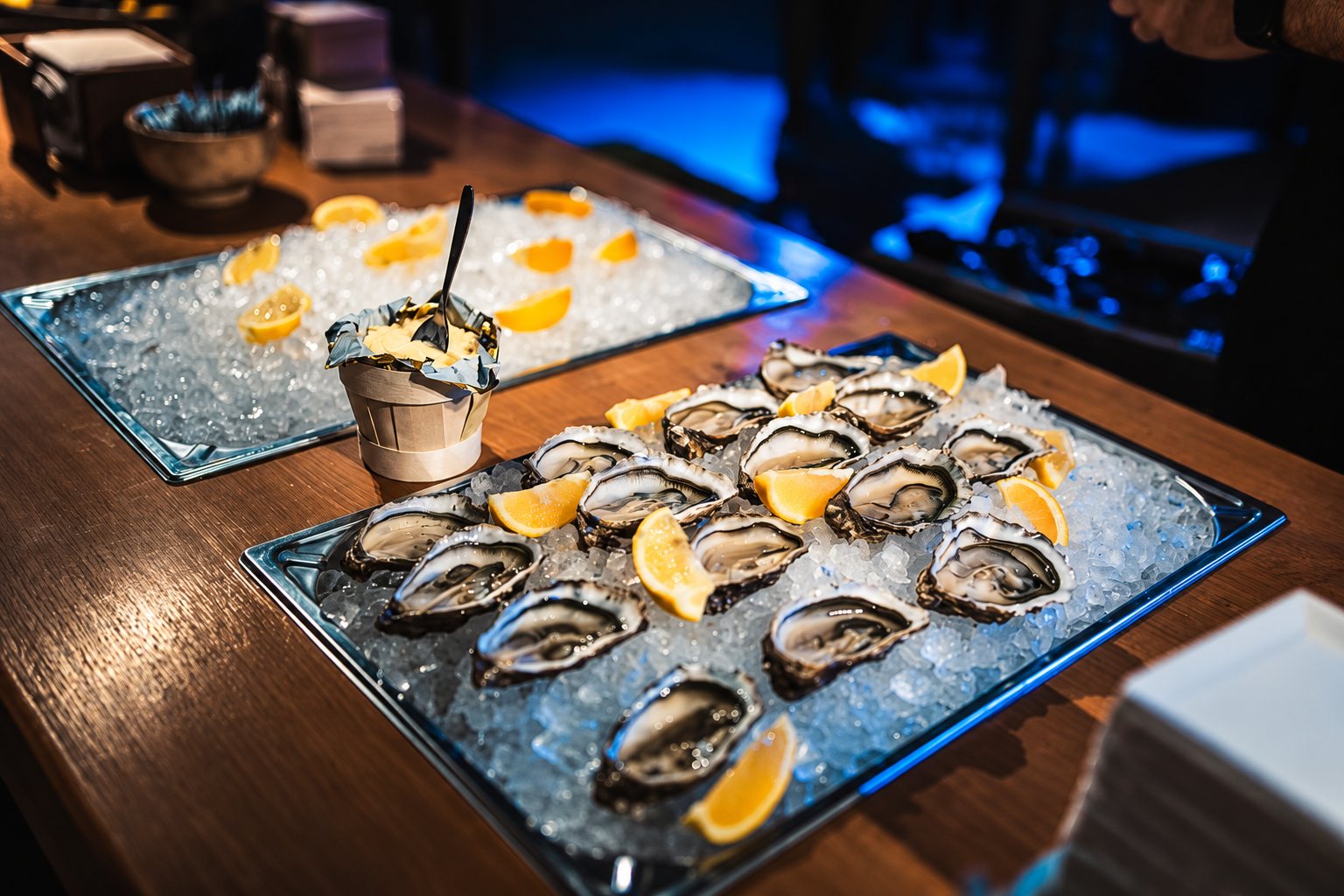 Deux plateaux métalliques remplis de glace pilée présentent des huîtres ouvertes et des quartiers de citron, posés sur un comptoir en bois sombre.