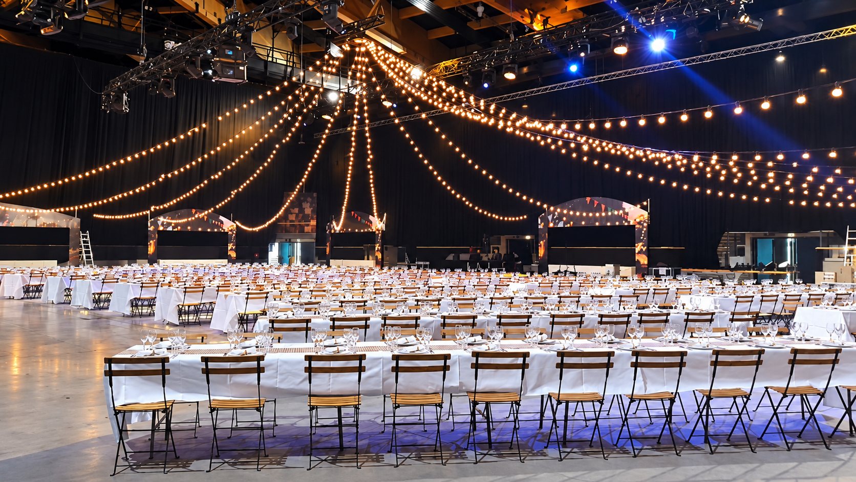 Une immense salle d'événement aménagée pour un banquet, avec de longues tables à nappes blanches, des chaises pliantes en bois et des guirlandes lumineuses suspendues au plafond.