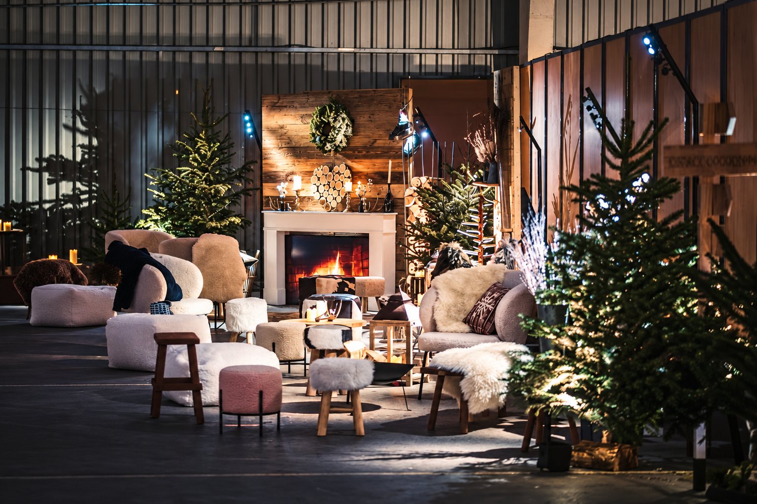 Un espace salon chaleureux dans un hangar, avec une cheminée allumée, des fauteuils et tabourets en fourrure et des sapins de Noël éclairés.