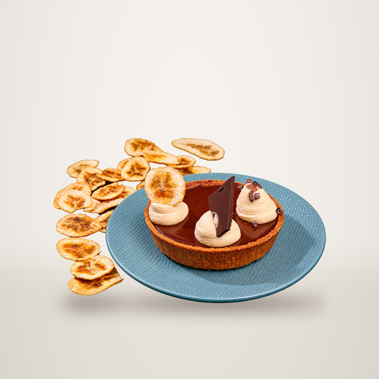 Une tartelette au chocolat garnie de chantilly, d'un carré de chocolat et de chips de banane, posée sur une assiette bleue texturée devant un fond noir.